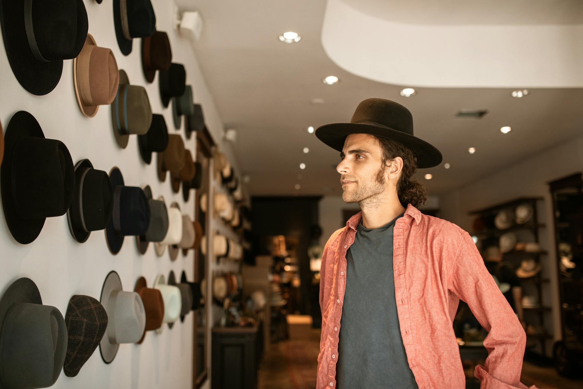 Man in a red shirt browsing fedora hats in a stylish, well-lit hat shop.